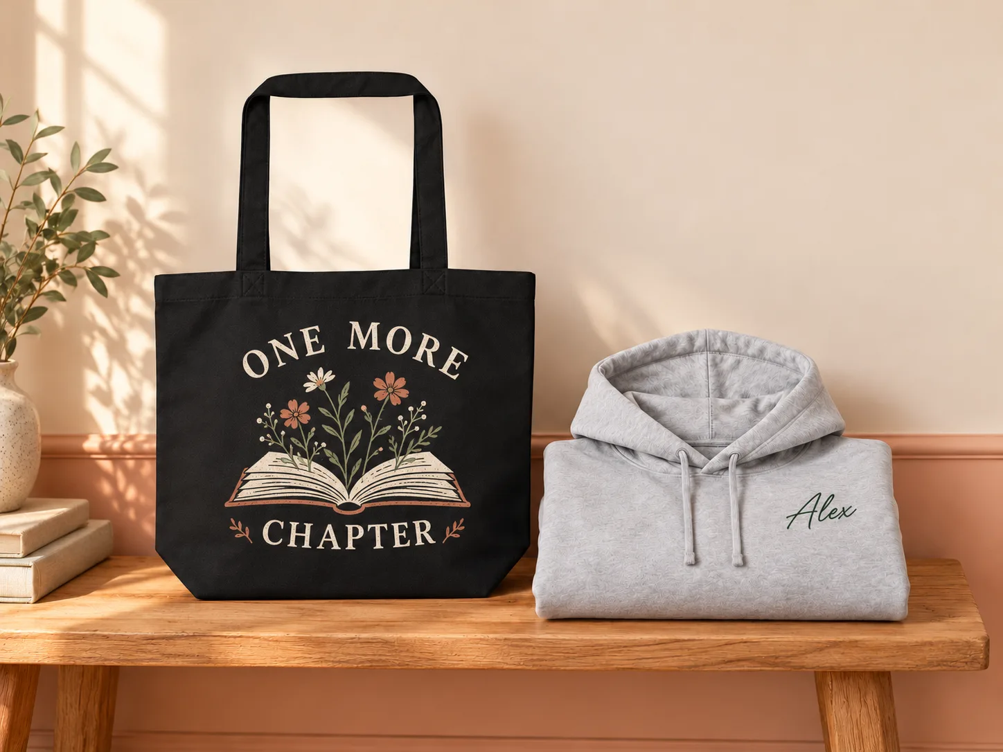 A black tote printed One More Chapter beside a grey hoodie embroidered with the name Alex, on a wooden bench